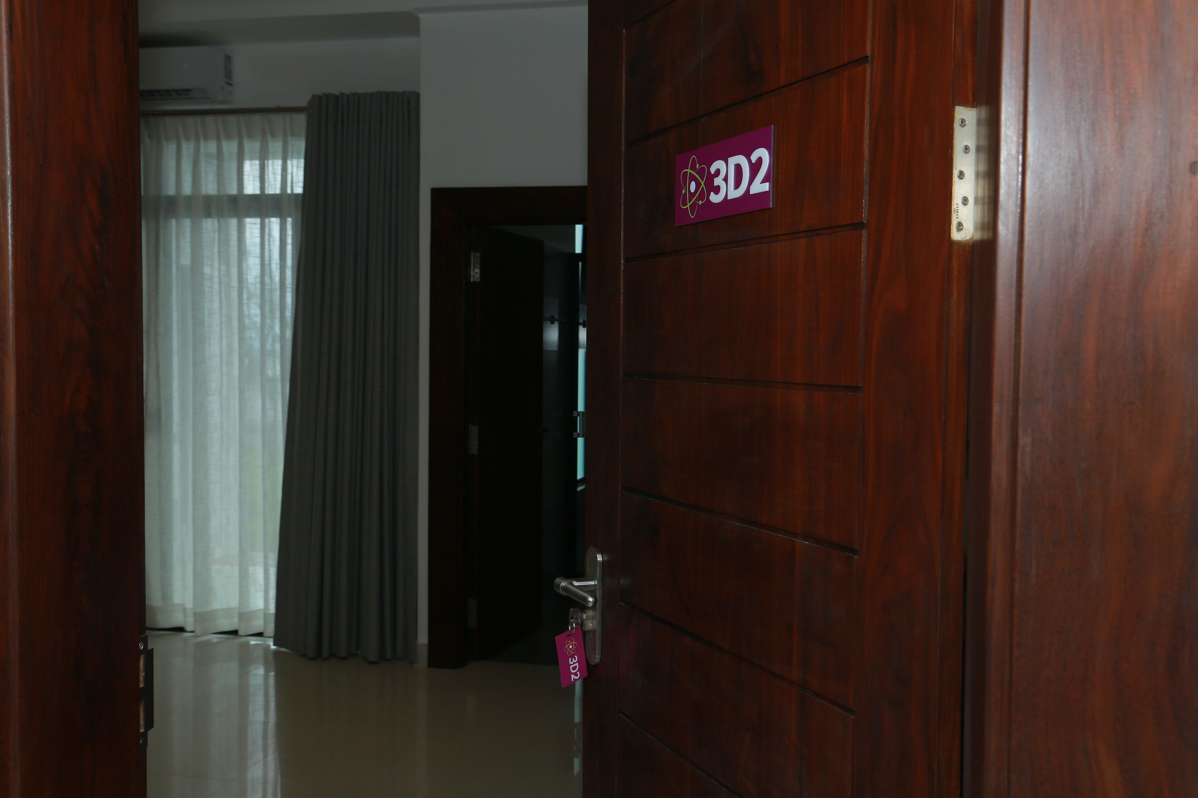 A wooden door with a 3D2 sign, a key in the lock, and a view of a room with a curtain and a door in the background.