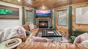 A cozy cabin interior with a stone fireplace, leather sofas, and a television mounted on the wall.