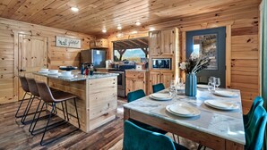 A wooden cabin interior with a kitchen, dining area, and a large window.