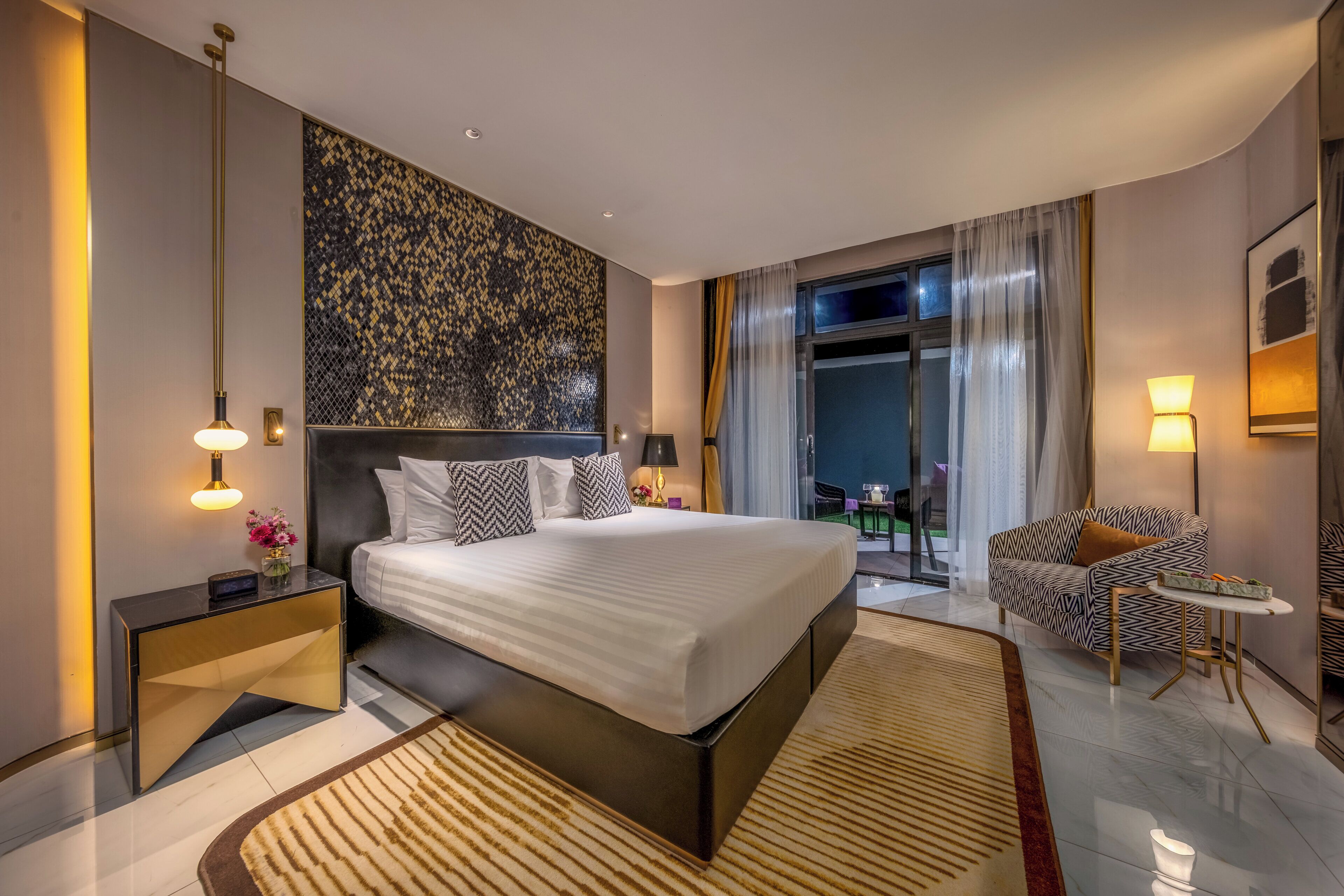 A modern hotel room with a large bed, a nightstand, a sofa, and a view of the outdoors.