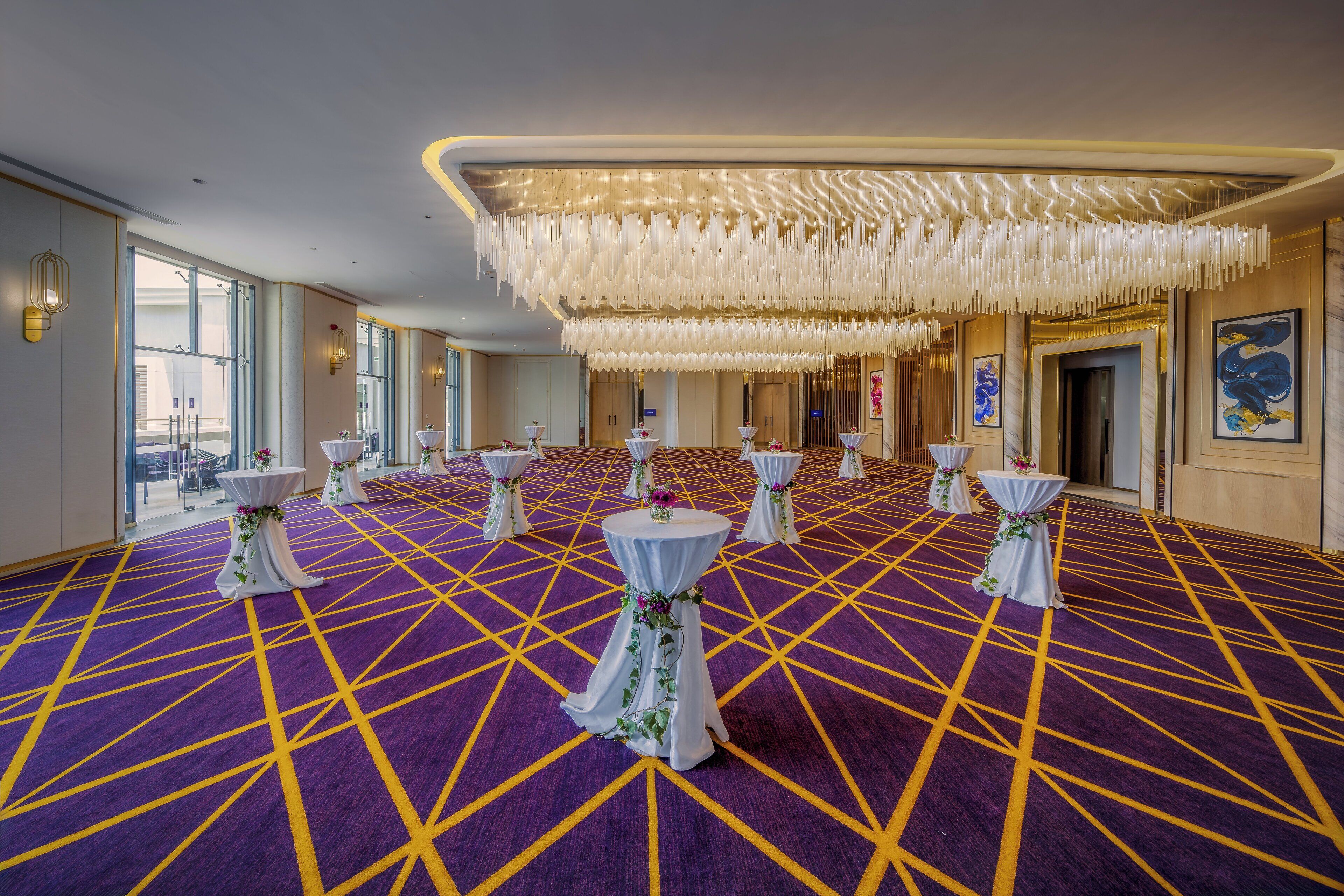 A spacious hall with a geometric patterned carpet, multiple round tables with white tablecloths and floral centerpieces, and a modern chandelier.
