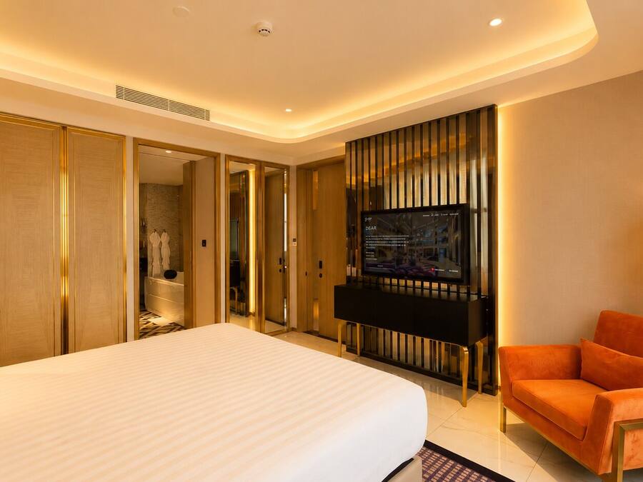 A hotel room with a large bed, a TV mounted on the wall, a seating area with a sofa, and a bathroom visible through an open door.