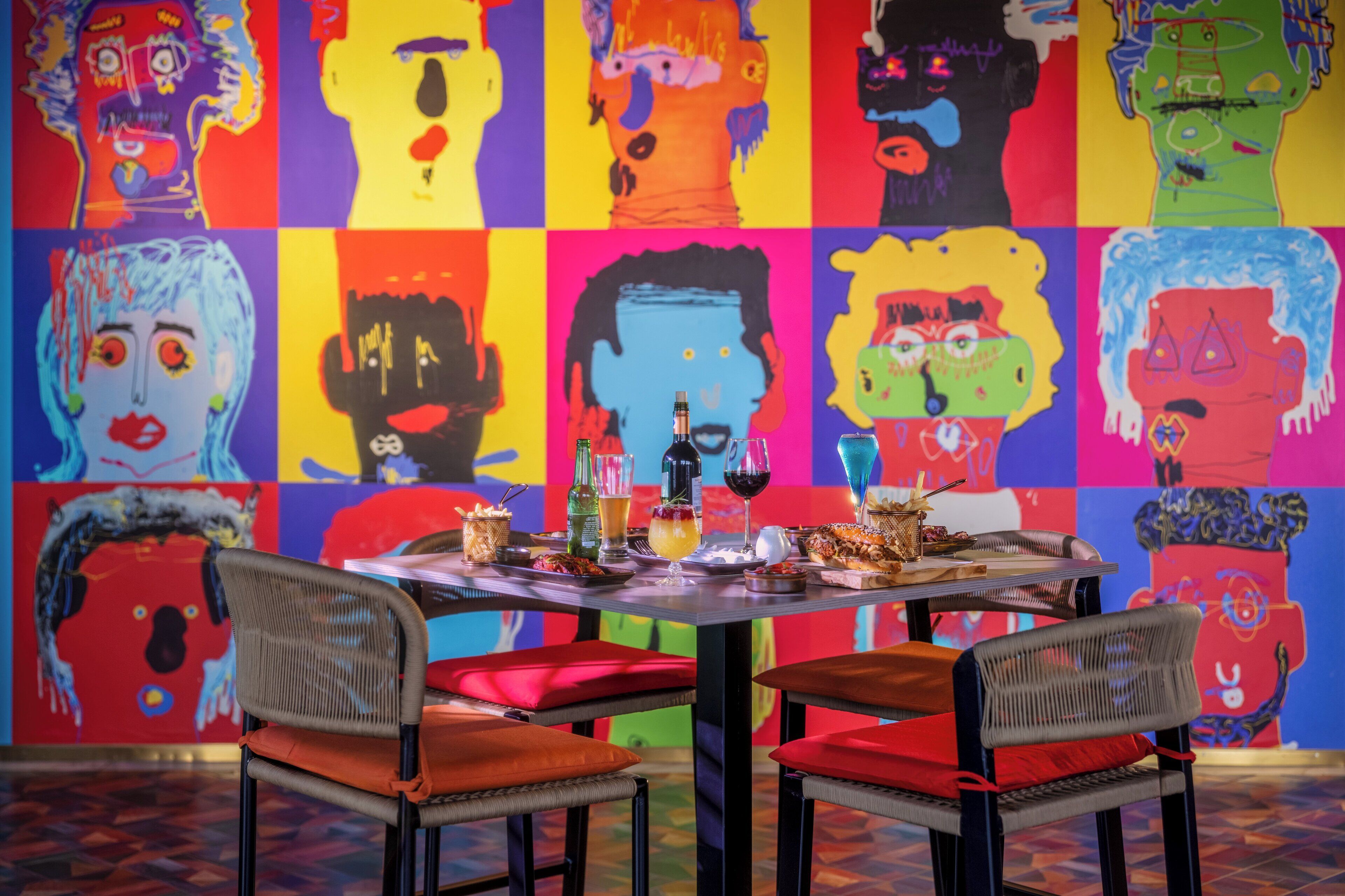 A dining area with a table set for two, chairs with red cushions, and a colorful abstract mural on the wall.