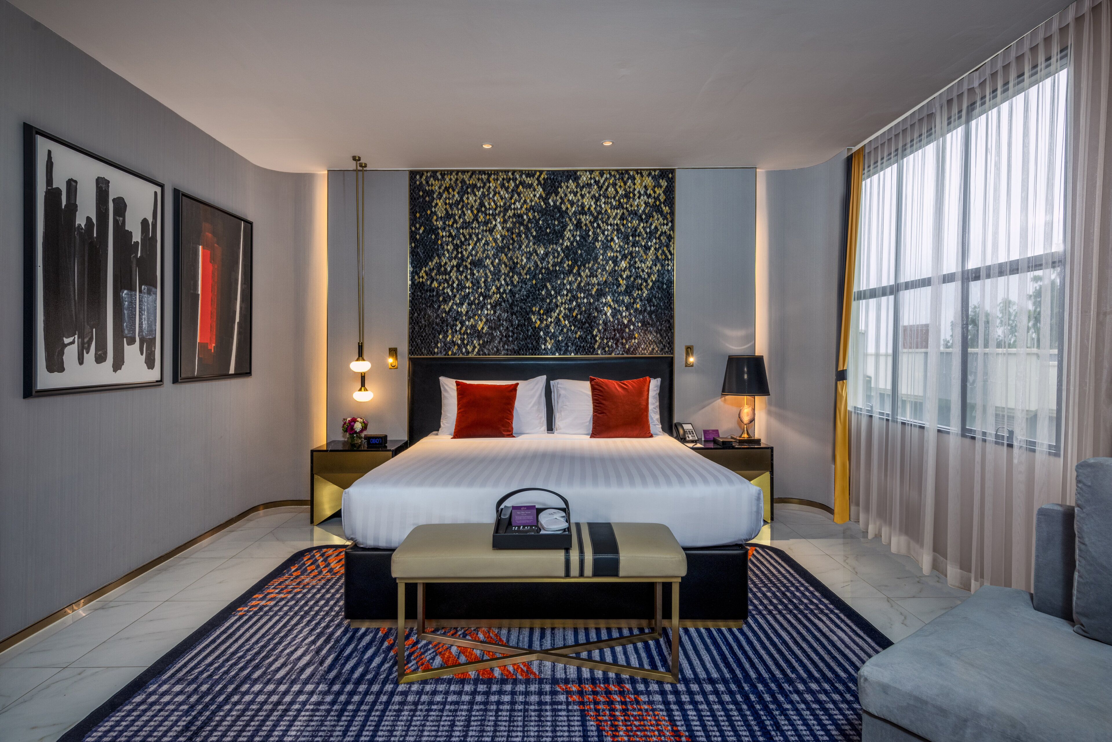 A modern hotel room with a large bed, a bench, a nightstand, and a view of the city through sheer curtains.
