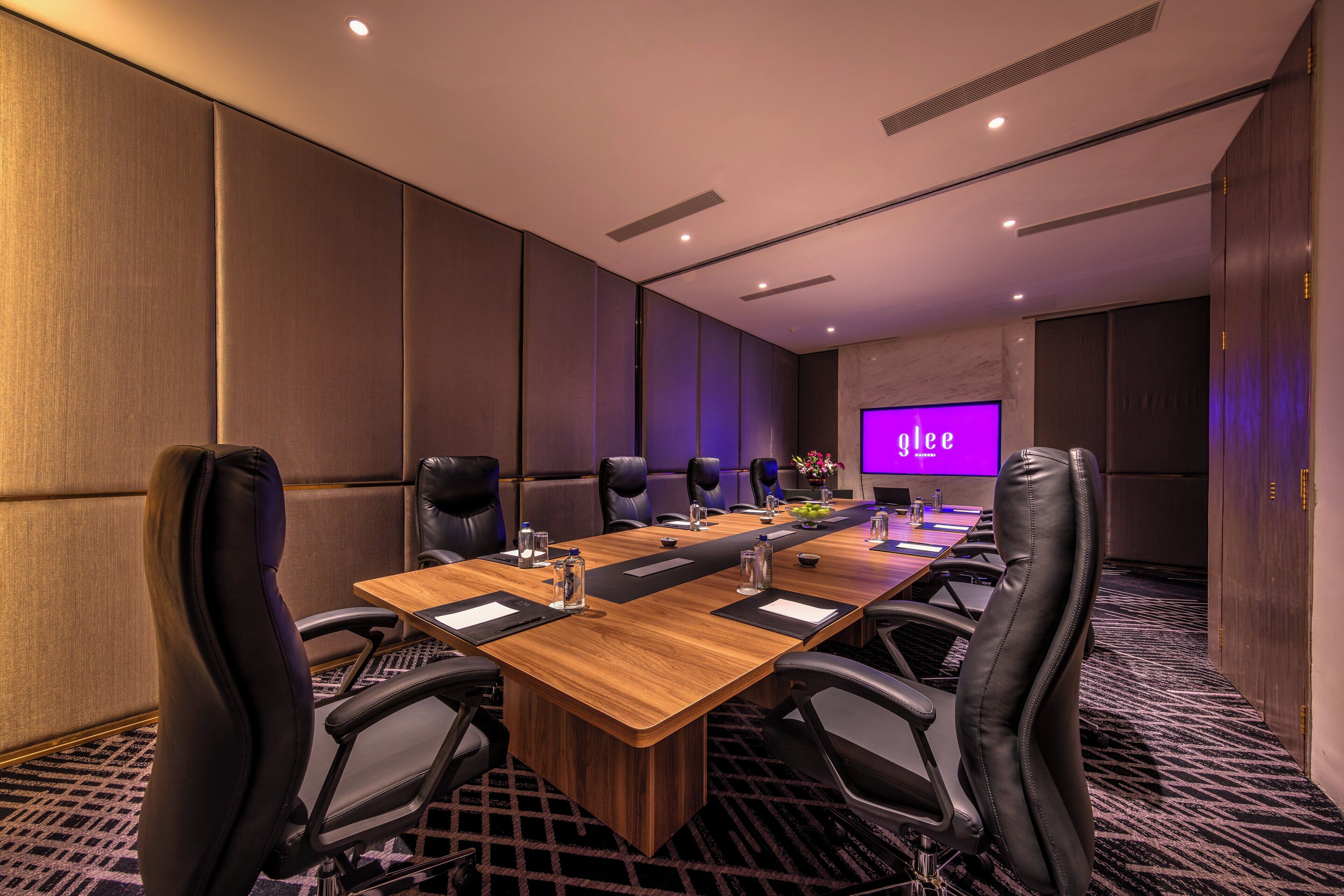 A conference room with a long wooden table, black leather chairs, a flat-screen TV, and a floral arrangement.