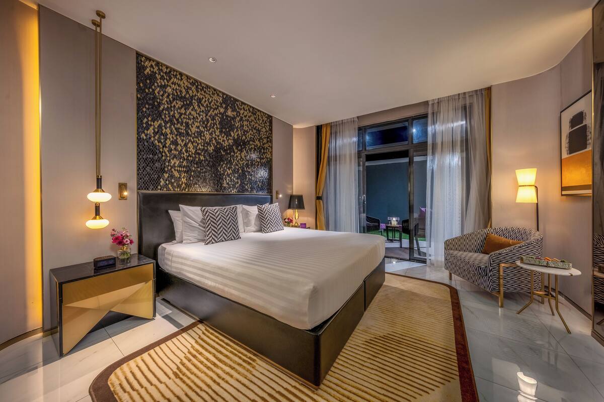 A modern hotel room with a large bed, a nightstand, a sofa, and a view of the outdoors.