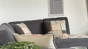 A grey sofa with multiple cushions, a woven plant pot, a wine glass, and a small table in a room with white paneled walls and a window with blinds. -  Beaches / Family Getaway / Amazing views / Golf / Wineries / Hot springs.
 


 (Rosebud)