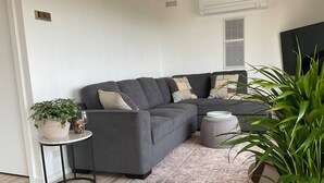 A modern living room with a grey sectional sofa, a small side table, a floor lamp, a large potted plant, and a patterned rug. -  Beaches / Family Getaway / Amazing views / Golf / Wineries / Hot springs.
 


 (Rosebud)