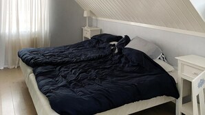 Een slaapkamer met een schuine kap, een bed met donkerblauwe dekbedovertrek, een nachtkastje met een lamp en een nachttafeltje.