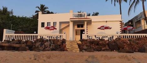 A two-story building with a balcony, a stone wall in front, and palm trees in the background.