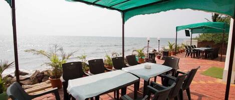 An outdoor seating area with tables and chairs under a green canopy, overlooking the sea.