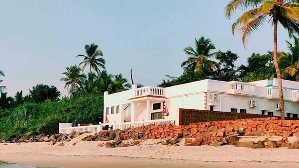A beachfront property with a white building, a stone wall, and palm trees. - White Seasons Resort And Spa (Malvan)