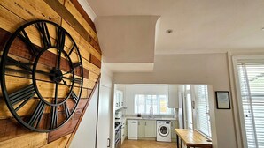 Une cuisine avec des meubles blancs, un lave-linge et un grand mur en bois orné d’une horloge.