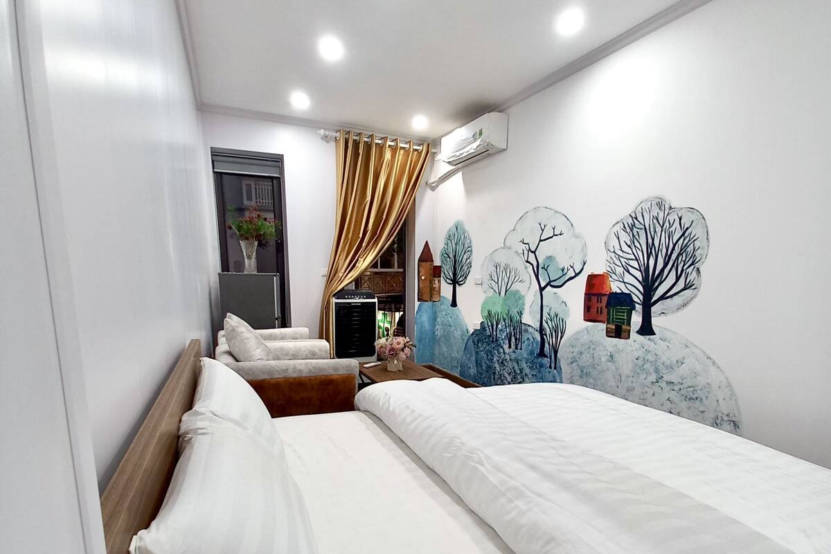 A hotel room with two beds, a window with a curtain, a wall-mounted air conditioner, and a mural of trees and houses.