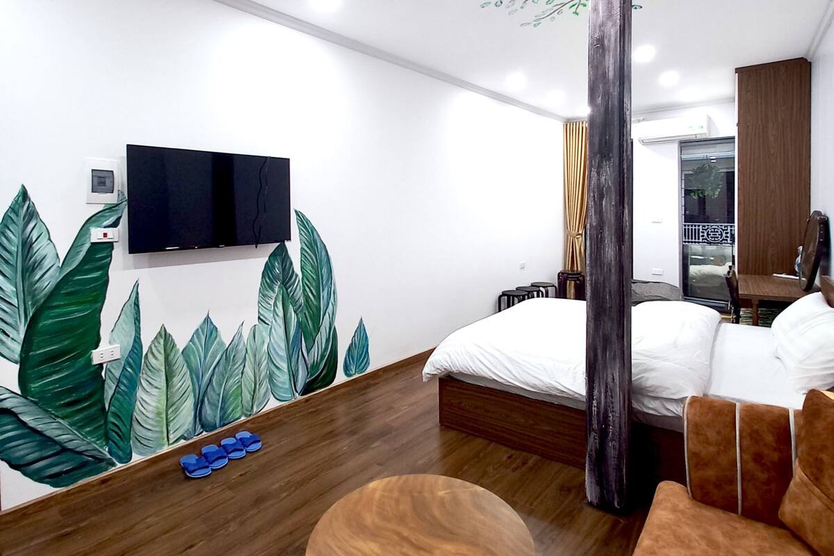 A modern hotel room with a large bed, a wooden coffee table, a TV mounted on the wall, and a large tree mural.