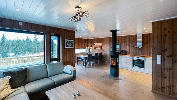 Interior modern amb estil de cabana de fusta, zona de menjador, cuina amb estufa de llenya i finestra gran amb vista a un paisatge nevós.