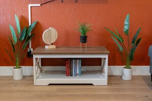 Una consola blanca con tapa de madera, flanqueada por dos macetas con hojas grandes, frente a una pared naranja.