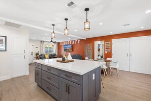 Una cocina moderna con una isla central, iluminación colgante y un comedor con mesa y sillas blancas.