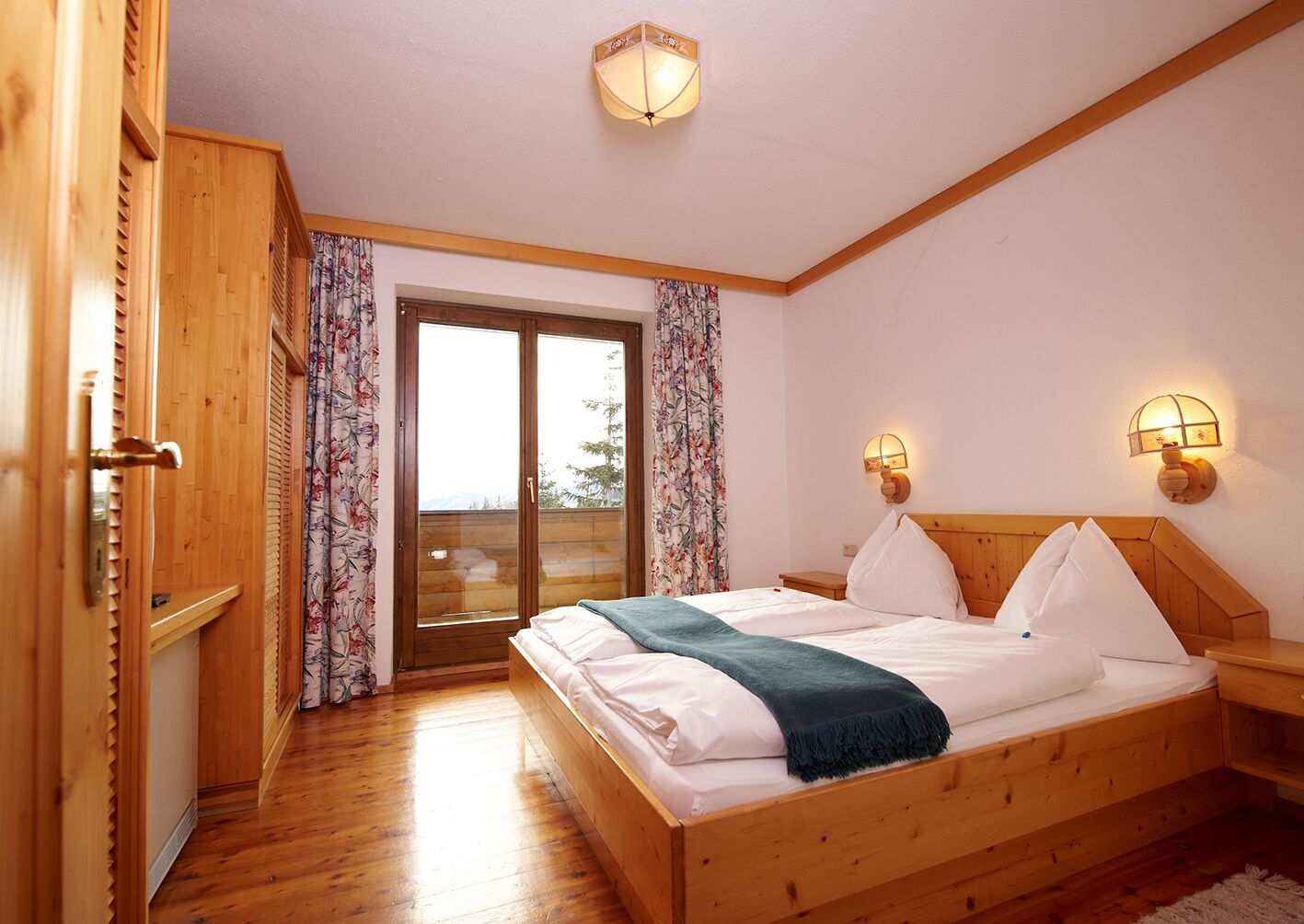Un dormitorio con estructura de madera, una cama, dos lámparas fijadas a la pared y un ventanal corredero a un balcón.