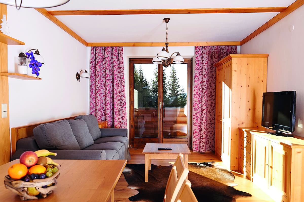 A cozy living room with a sofa, a dining table with fruit, a TV, and a view of trees through a glass door.