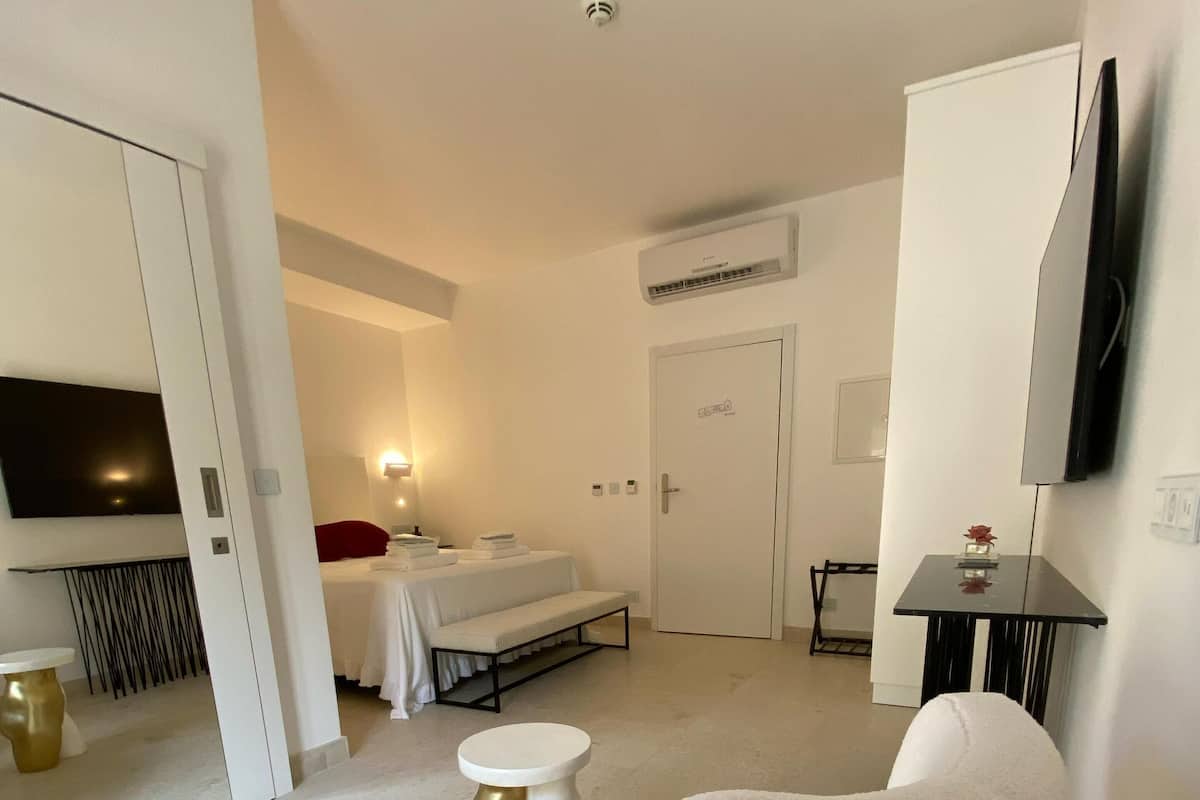 A modern hotel room with a bed, a sitting area with ottomans, a TV mounted on the wall, and a door with a sign.