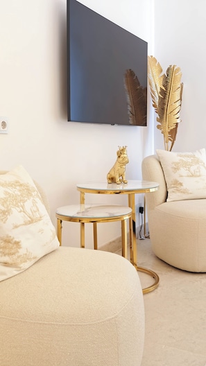 Uma sala de estar moderna com um sofá, uma pequena mesa redonda com uma figurinha de cachorro decorativa, uma televisão na parede e uma pena dourada decorativa.