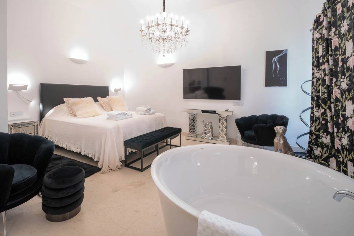 A modern bedroom with a large bed, a bathtub, a TV, and a chandelier.