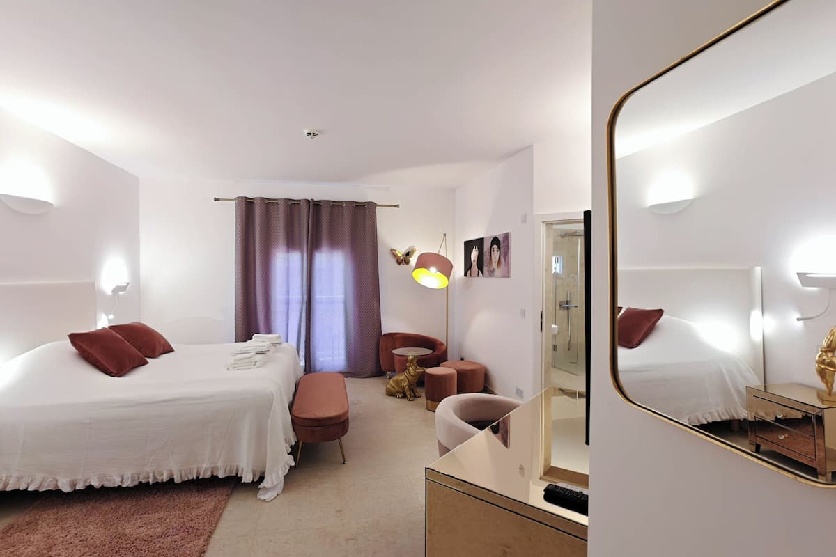 A modern hotel room with a large bed, a sitting area, a mirror, and a shower.