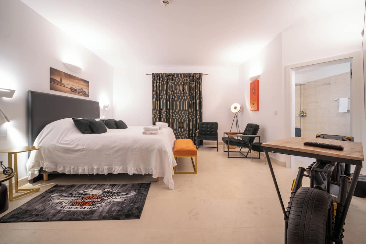 A modern hotel room with a bed, bedside table, chair, desk, and a wheelchair.