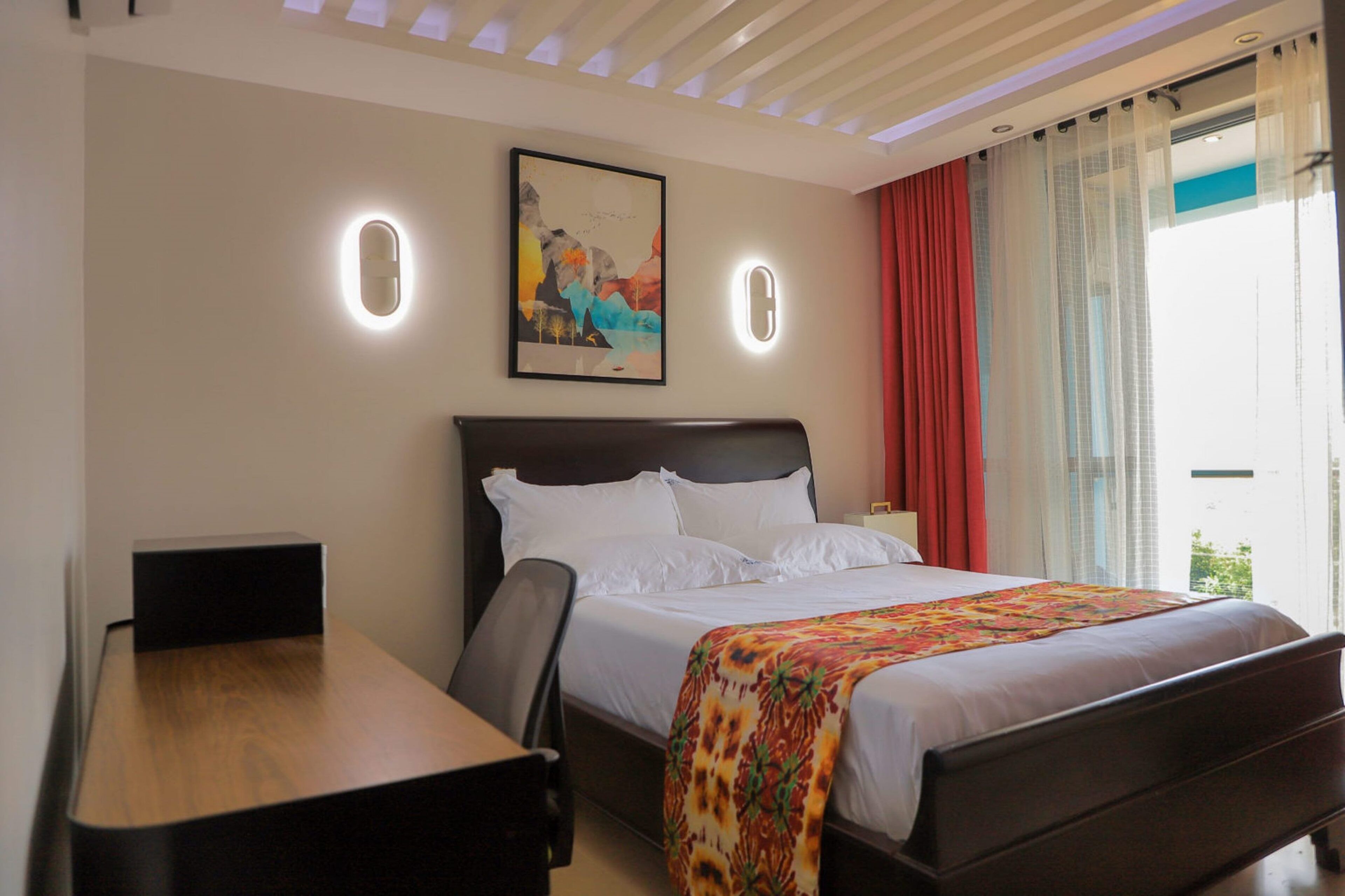 A hotel room with a bed, a desk, and a painting on the wall.