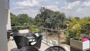 A balcony with wicker furniture, a glass railing, and a view of trees and a building in the background. - Le tsuba Grand Hotel (Arua)