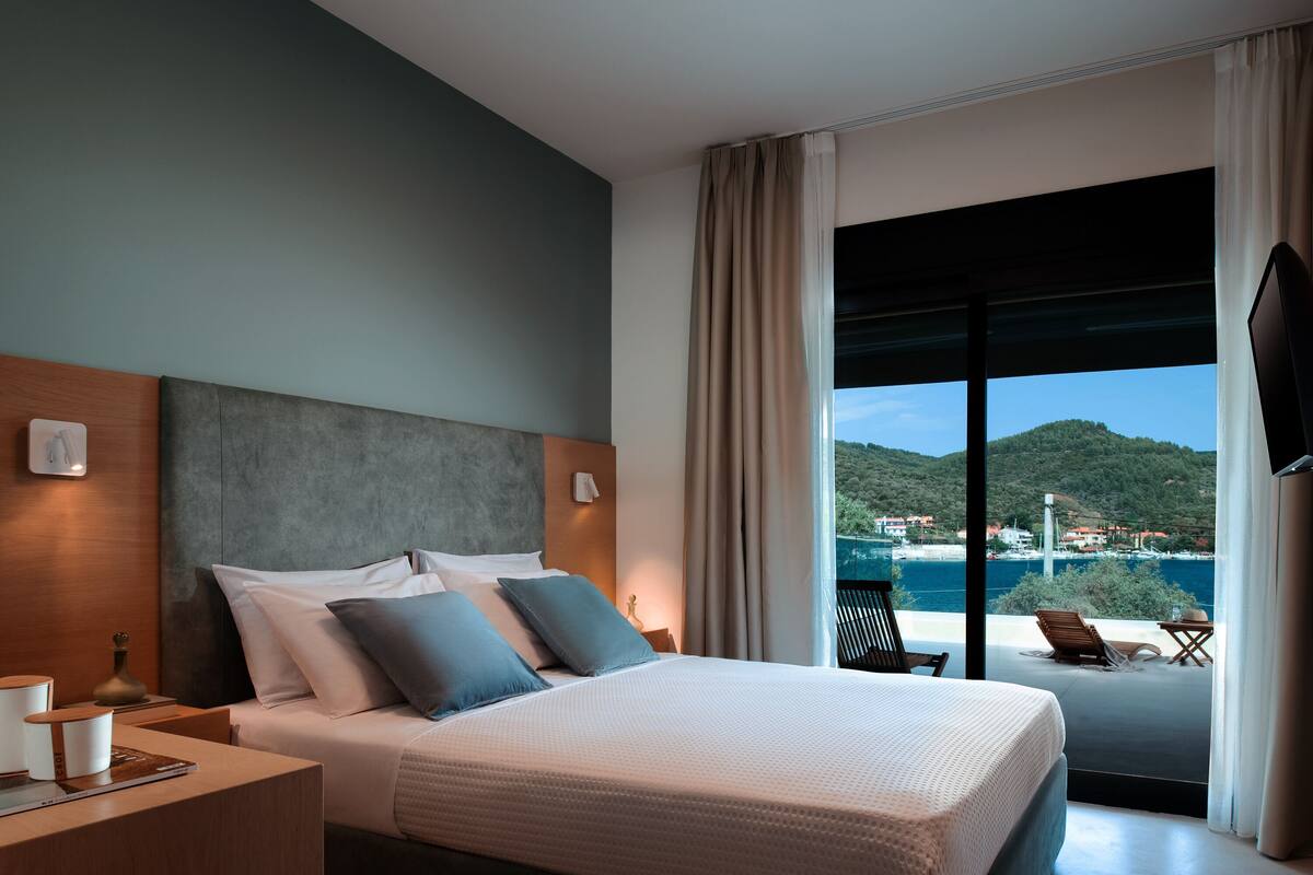 A hotel room with a large bed, a nightstand, a TV, and a balcony with a view of the sea and hills.