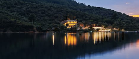 A lakeside property with a large house surrounded by dense forest, reflecting on the calm water.