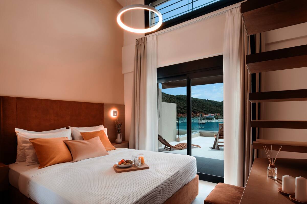 A modern hotel room with a large bed, a breakfast tray, a view of a pool and mountains, and a balcony with a chair.