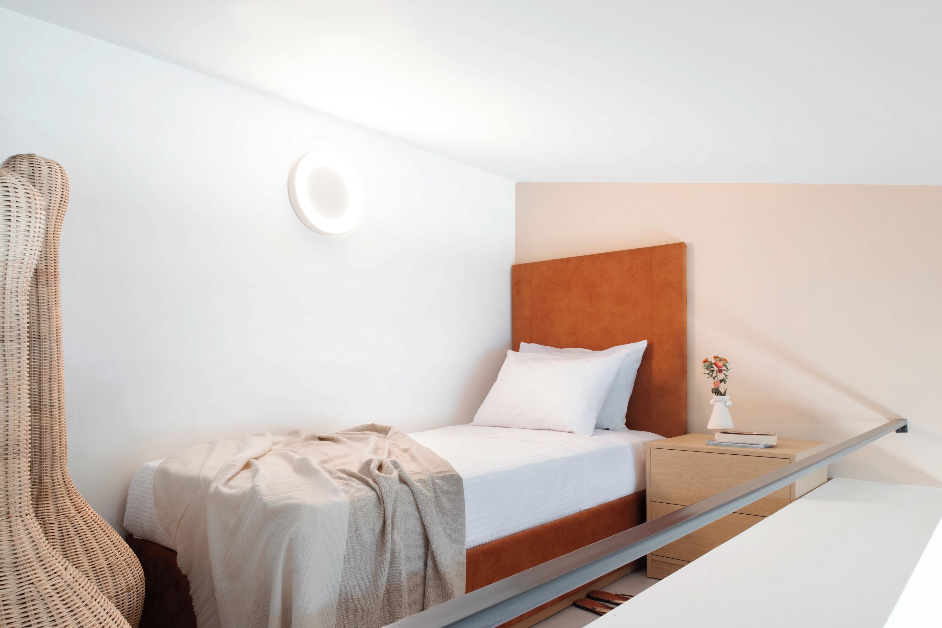 Une chambre d’hôtel moderne avec un lit simple, une table de chevet, une petite table et une chaise en osier.