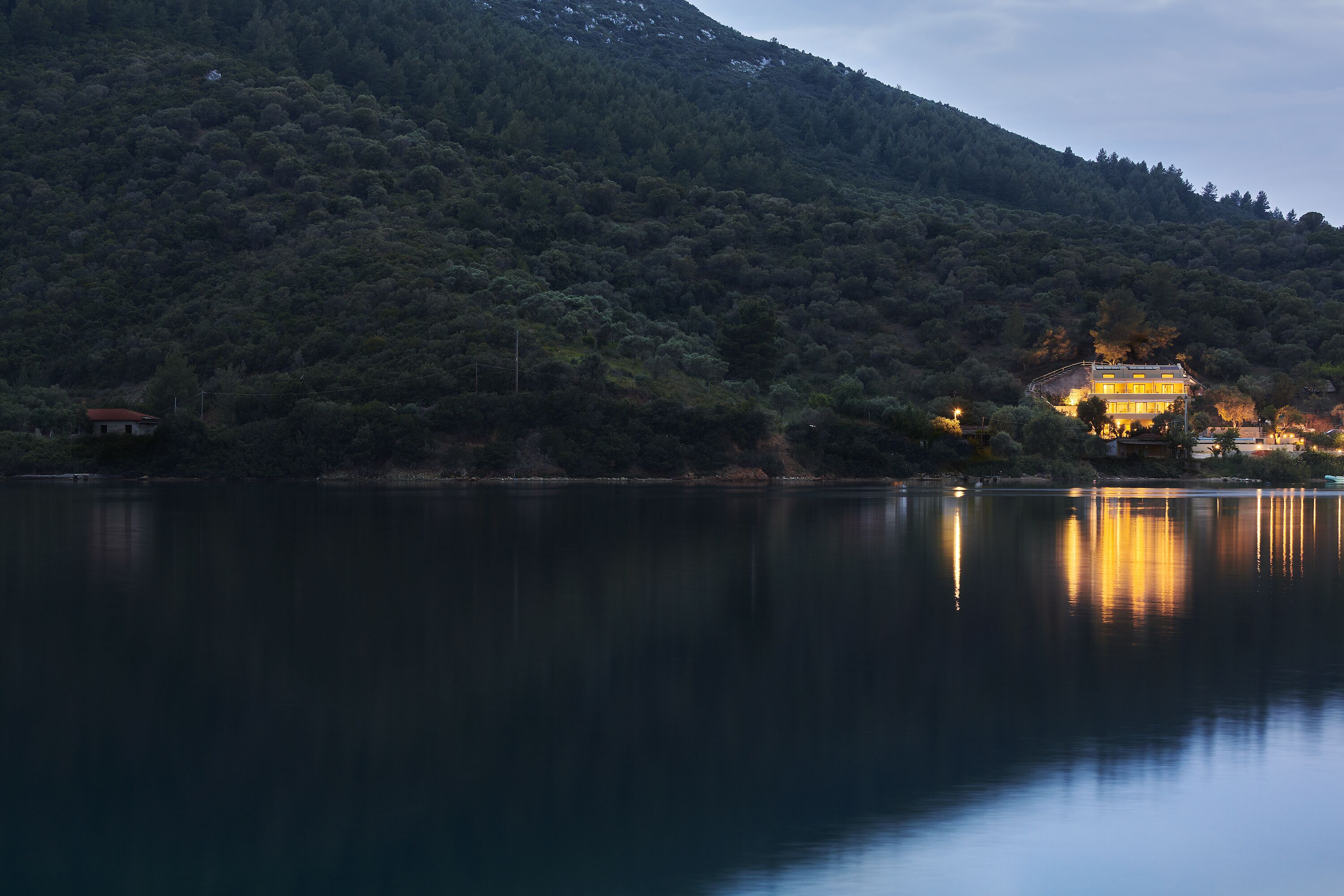 A house with a visible balcony, surrounded by a forested hillside and a calm lake.