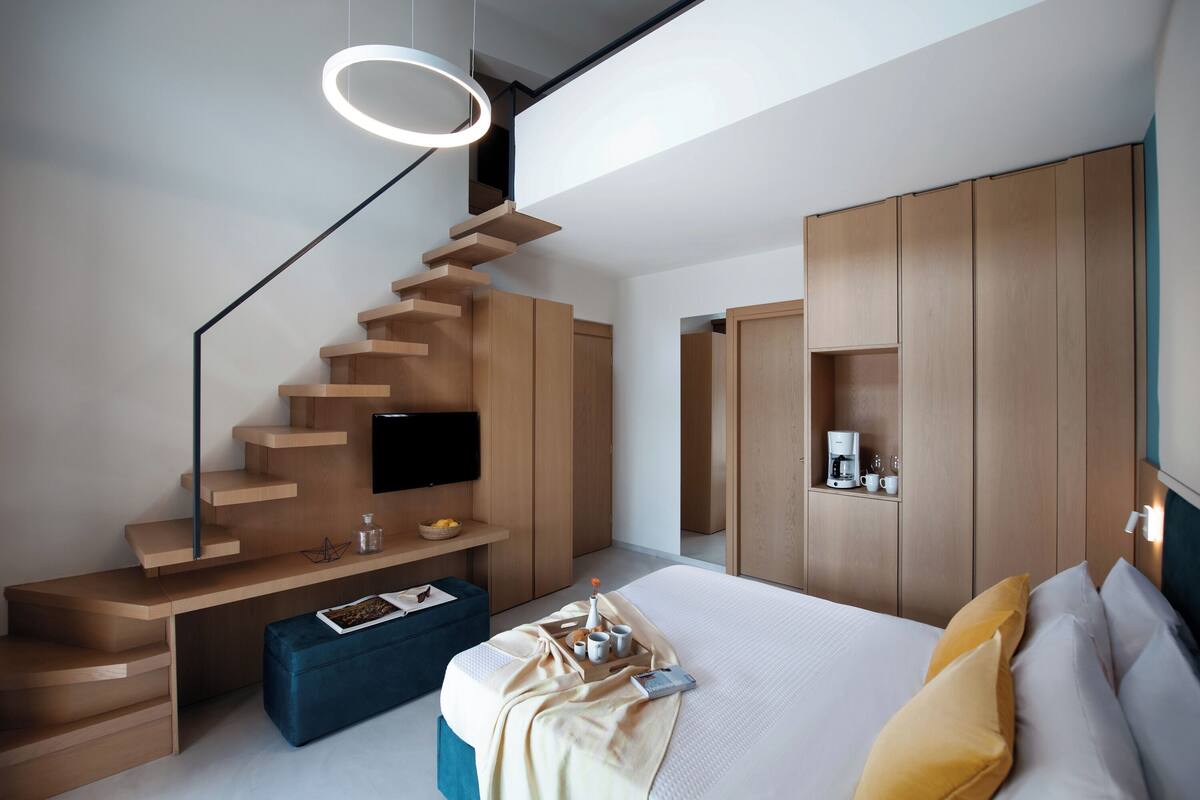 A modern bedroom with a wooden staircase, a flat-screen TV, a bedside table with a tray, and a large bed with pillows.