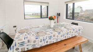 A dining area with a table set for four, two windows with views of the sea and buildings, and a vase with flowers on the table. - Old Field House (Templetown)