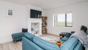 A modern living room with teal sofas, a flat-screen TV, a bookshelf, and a large window with a view of the sea. - Old Field House (Templetown)