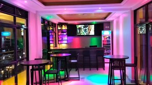 Zona de bar con varias mesas y sillas, una pantalla de televisión e iluminación colorida.
