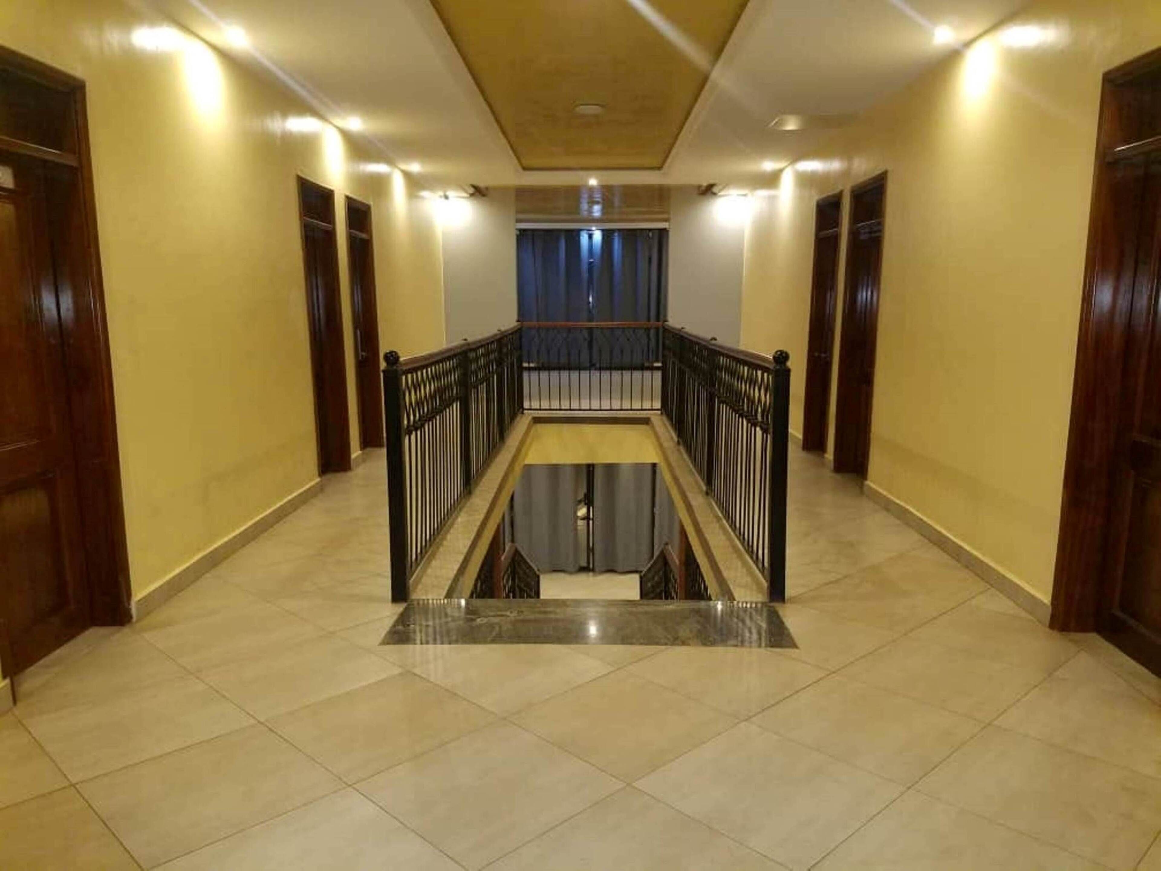 A hotel corridor with a central glass elevator, wooden doors, and a tiled floor.
