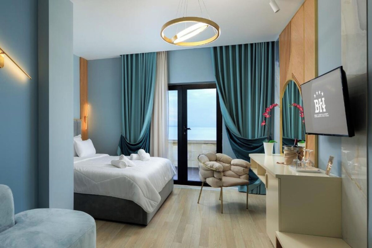 A modern hotel room with a bed, a desk, a chair, a TV, and a view of the sea.