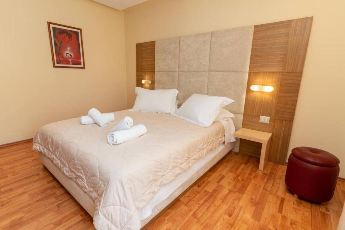 Una cama bien hecha con sábanas y almohadas blancas, cabecera de madera, mesita de noche con lámpara y un otoman rojo.