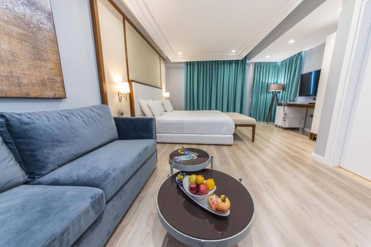 Una habitación de hotel con una cama, un sofá, una mesita redonda con fruta, una lámpara y un televisor.