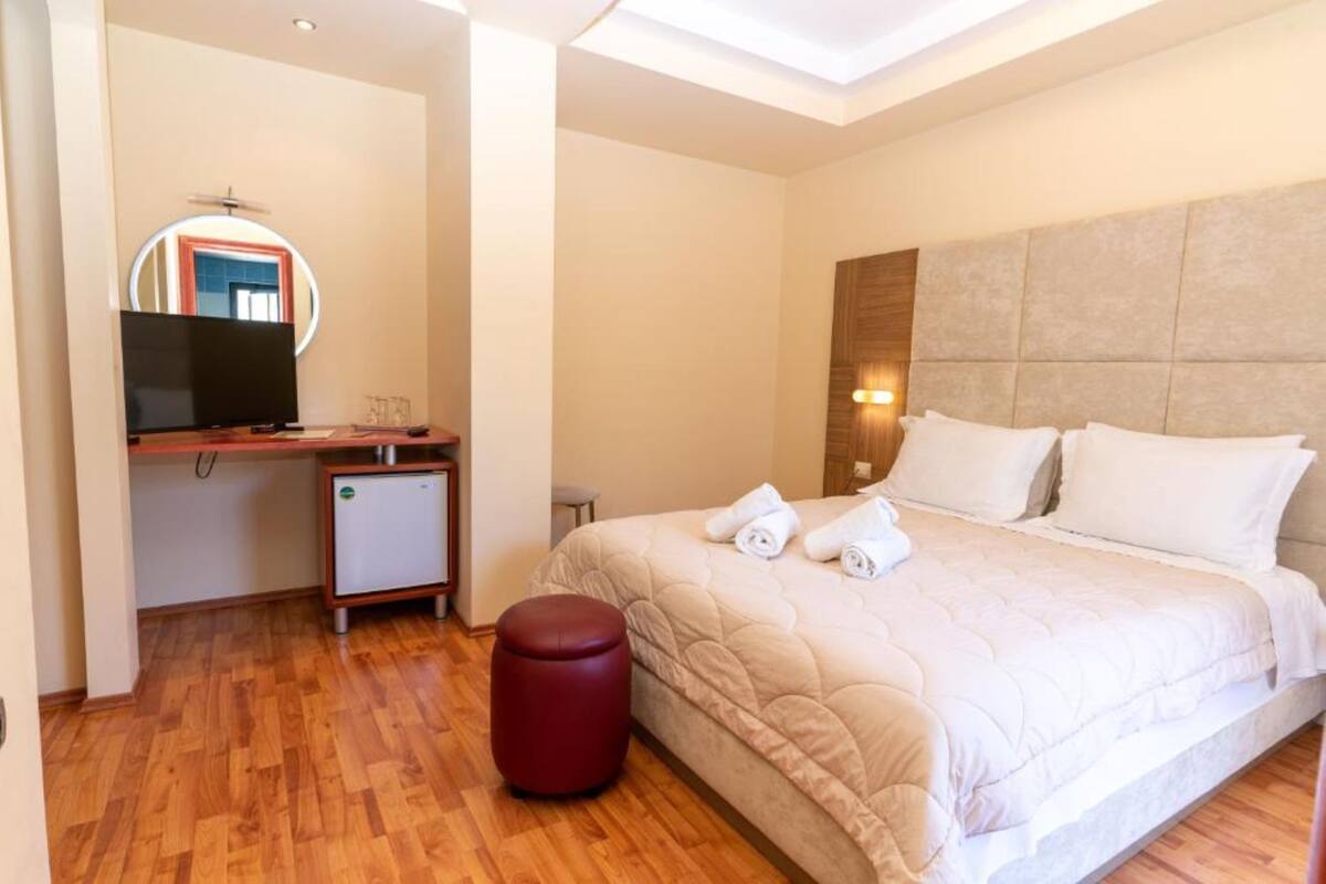 Una habitación de hotel con una cama, un escritorio con televisión, un refrigerador pequeño y un taburete rojo.
