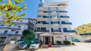 A multi-story hotel with a modern design, featuring balconies on each floor, a prominent entrance, and a parking area with several cars. - Hotel Brilant (Sarandë)