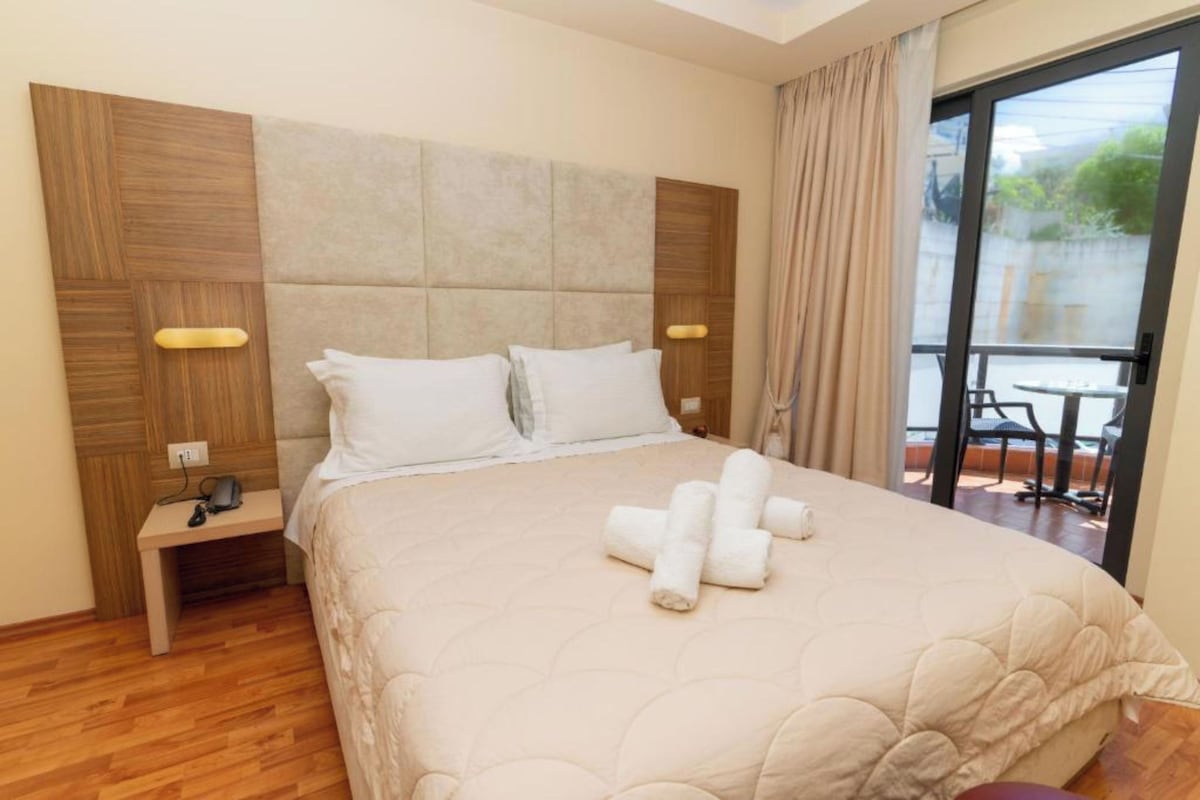 A hotel room with a large bed, a nightstand with a phone, and a view of the outside balcony.