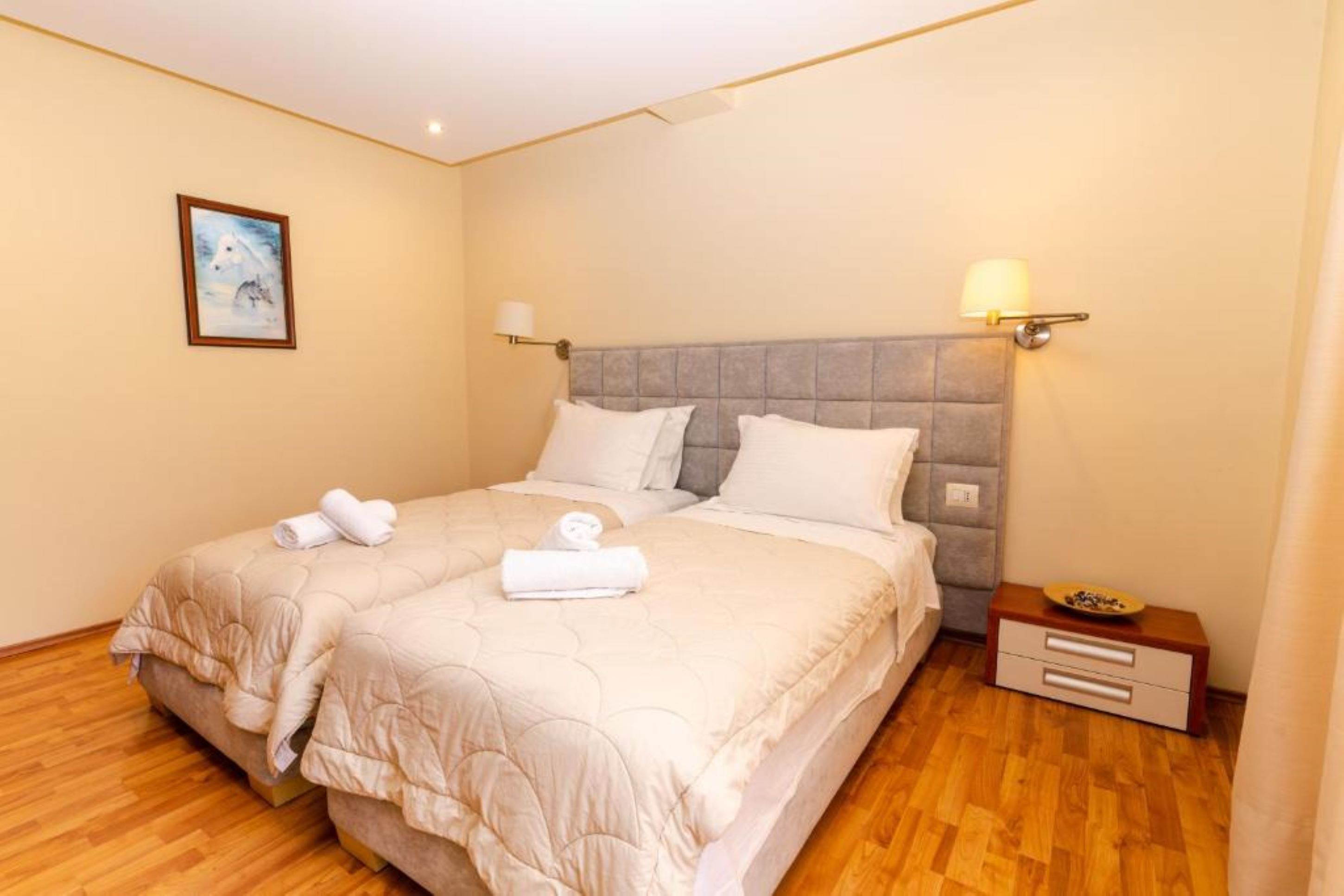 A double bed with a grey headboard, two bedside lamps, a small bedside table with a bowl, and a framed picture on the wall.