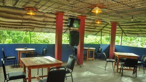 An indoor dining area with tables and chairs, a television mounted on a pillar, and a view of greenery outside.