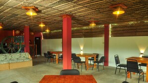 A spacious indoor area with wooden tables and chairs, a large green decorative wheel, and a thatched ceiling.